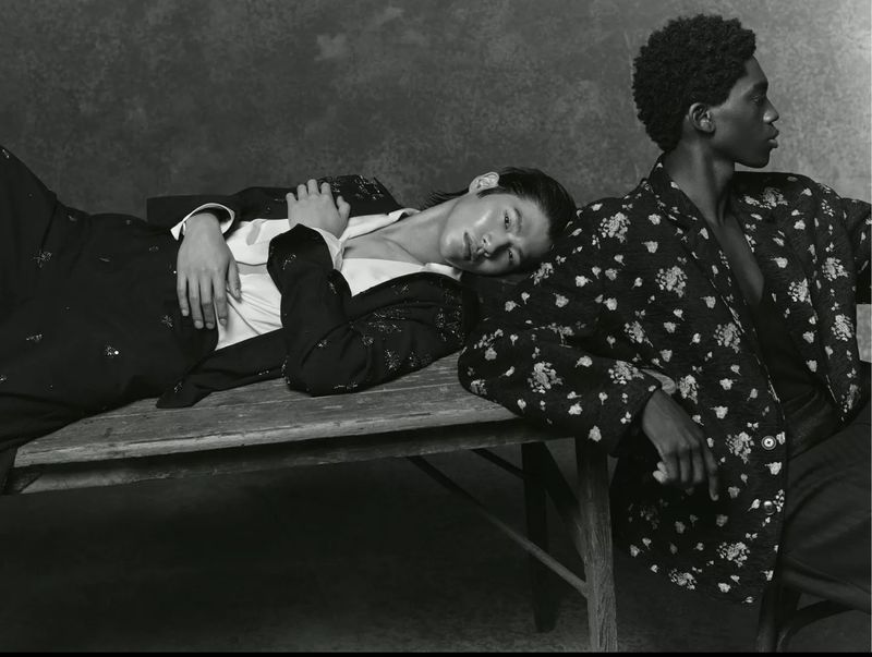 Two male fashion models pose indoors on a wooden surface wearing dark blazers and dress pants in black and white studio photography.