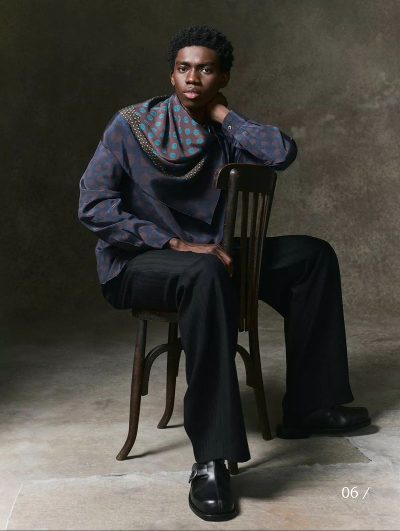 Portrait of an adult male sitting on a wooden chair in a studio setting wearing a blue patterned shirt, black pants, and black buckle shoes.