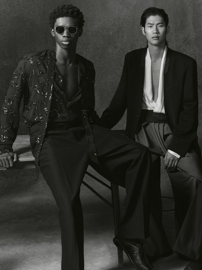 Two fashion models pose indoors against a grey backdrop, wearing black and white outfits, seated closely for a studio portrait.