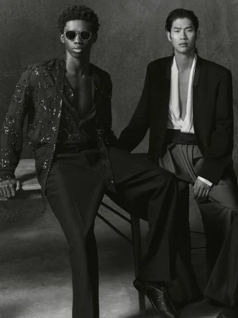 Two fashion models pose indoors against a grey backdrop, wearing black and white outfits, seated closely for a studio portrait.