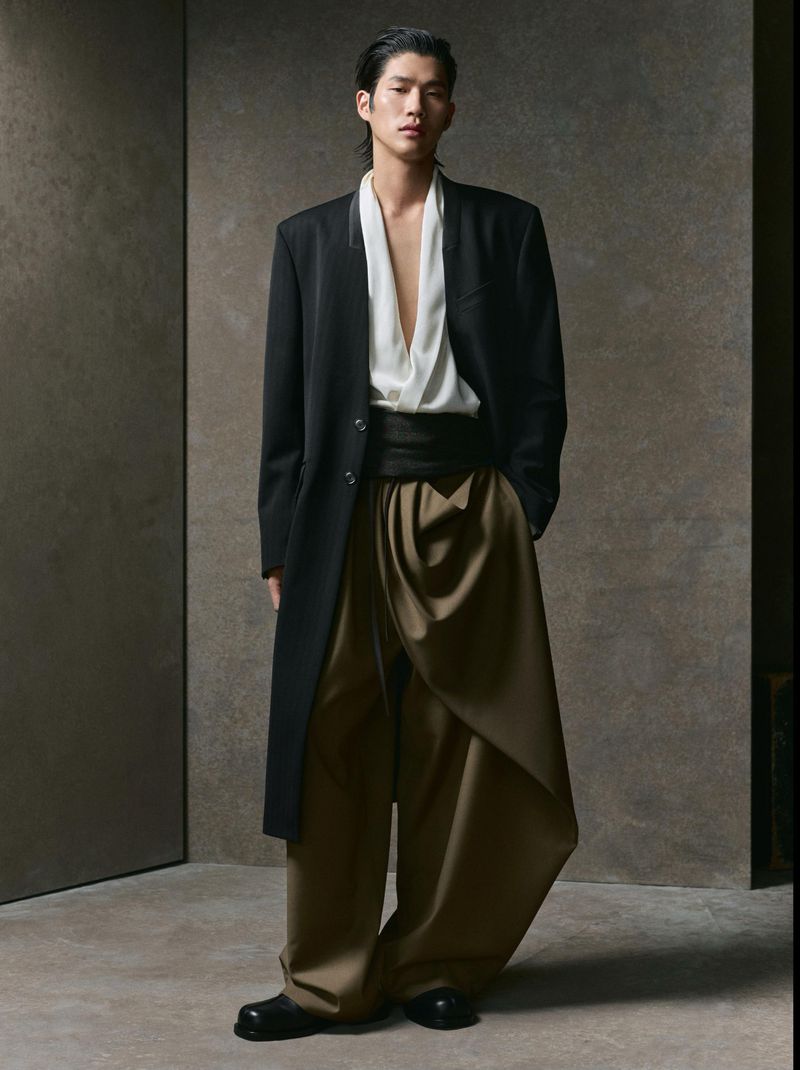 Asian man in long black coat and loose pants poses indoors against neutral backdrop in a studio setting for fashion editorial.