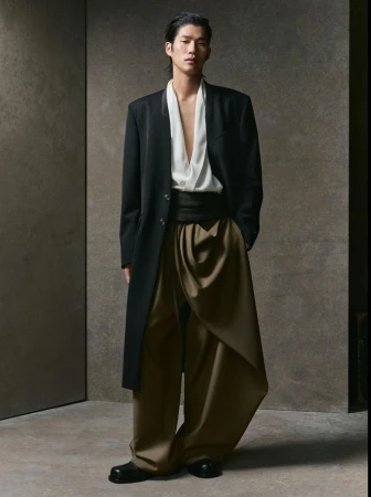 Asian man in long black coat and loose pants poses indoors against neutral backdrop in a studio setting for fashion editorial.