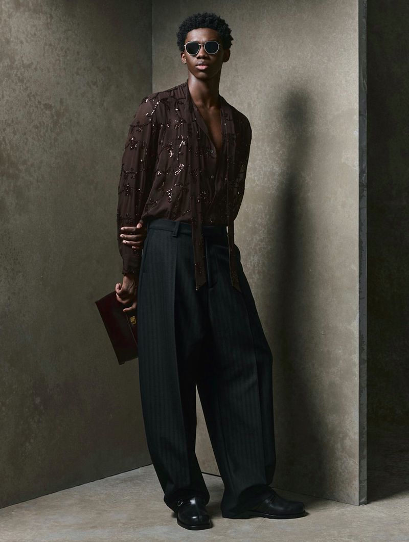 An adult Black man in a brown semi-sheer top and pinstripe pants poses in a studio with a dark book, complemented by dark sunglasses and shoes.