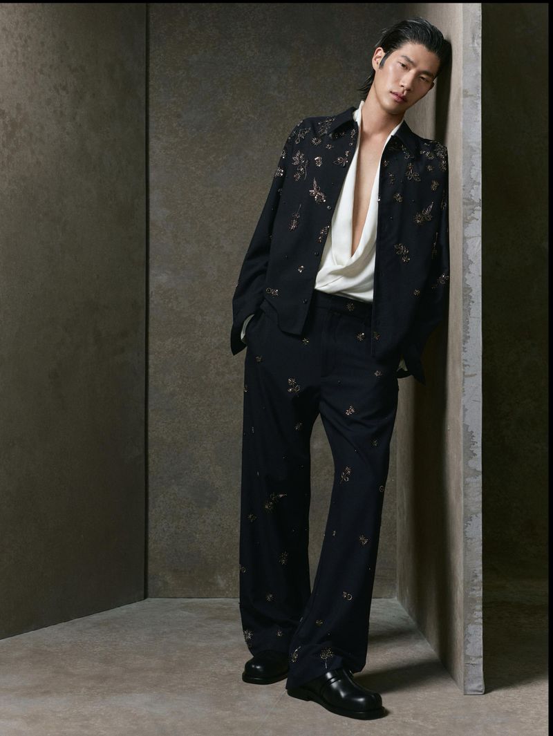 Model leaning against a dark textured wall wearing a black patterned shirt and trousers for Zara