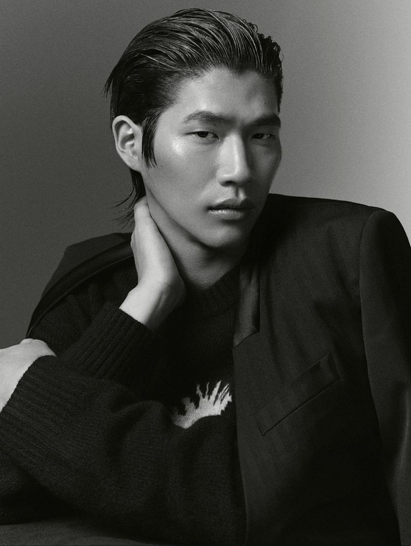 Portrait of an Asian male model with slicked-back hair. He is wearing a black jacket over a patterned sweater in a photo studio setting.
