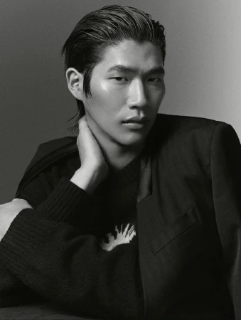 Portrait of an Asian male model with slicked-back hair. He is wearing a black jacket over a patterned sweater in a photo studio setting.