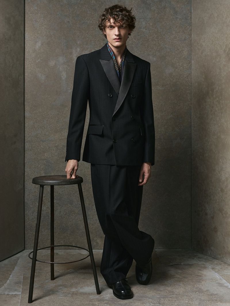 A young adult man wearing a black suit with a scarf stands in front of a neutral background, leaning on a stool in a studio shot.