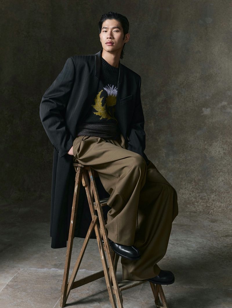 A single adult male sits on a wooden stool, wearing a black coat, a printed sweatshirt, and loose brown trousers in a studio setting.