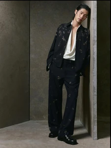Model leaning against a dark textured wall wearing a black patterned shirt and trousers for Zara