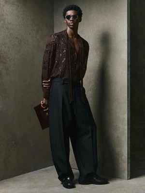 Model wearing dark sunglasses, brown sheer shirt, and pleated black trousers in front of a textured concrete studio corner.