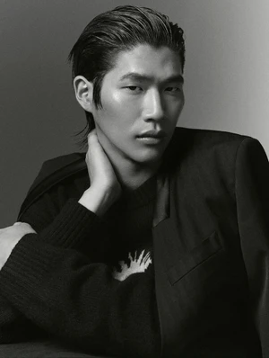 Portrait of a man with slicked-back dark hair wearing a black jacket and sweater against a grey studio backdrop.