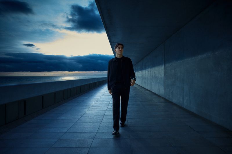 A man in dark clothing walks along a concrete walkway towards the camera, with a cloudy sky and ocean visible in the background.