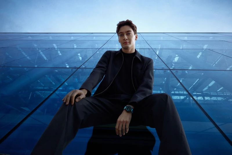 Man in a dark suit seated outdoors against a geometric blue glass building, captured from a low angle emphasizing the building's structure.