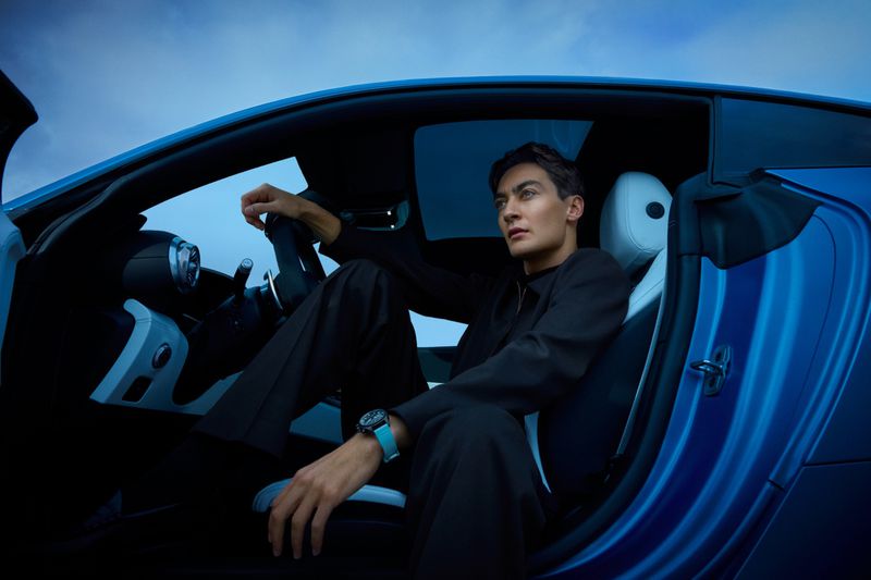 Adult male in black suit sitting in blue sports car with door open looking toward sky, showing wristwatch with turquoise band.