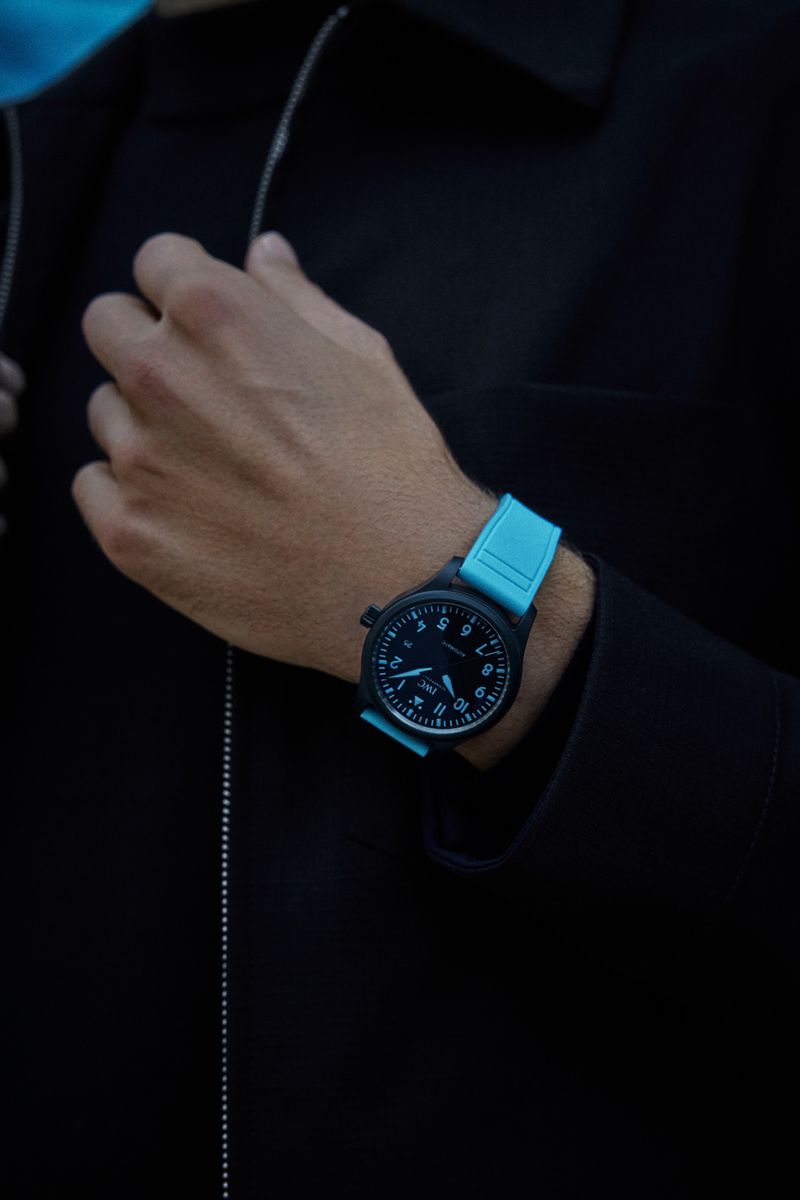 Close-up studio shot of an adult male's wrist wearing a black and light blue wristwatch with a black jacket and silver chain detail.