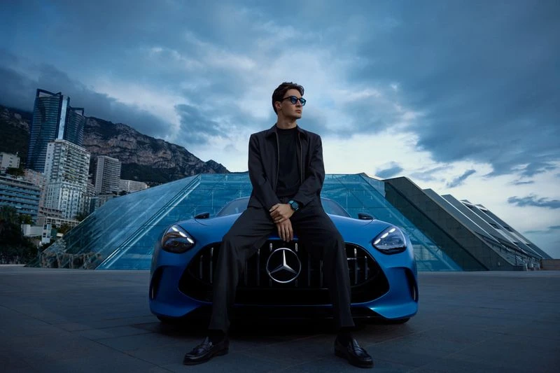 A man sits on the hood of a blue Mercedes in an urban setting with tall buildings, wearing sunglasses, a black suit, and black leather shoes.