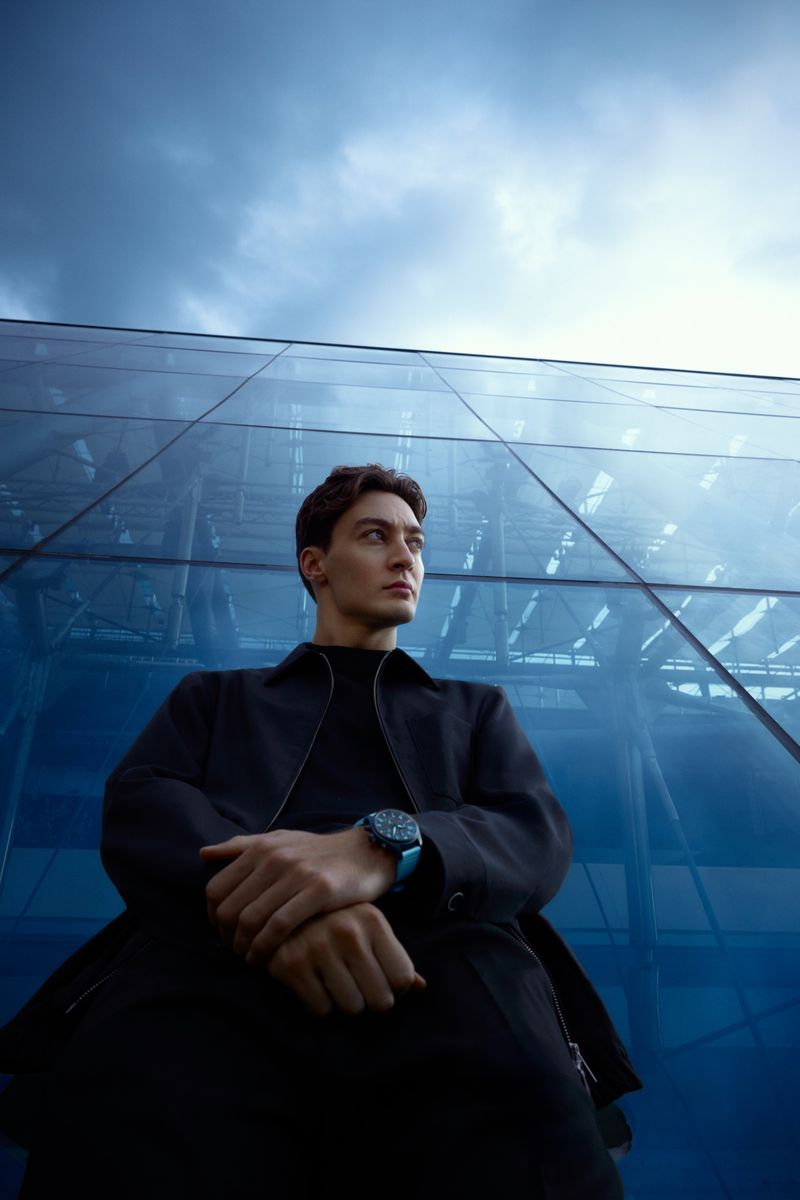 Low-angle portrait of a stylish adult male wearing a black overcoat and a watch, standing in front of a modern glass building.
