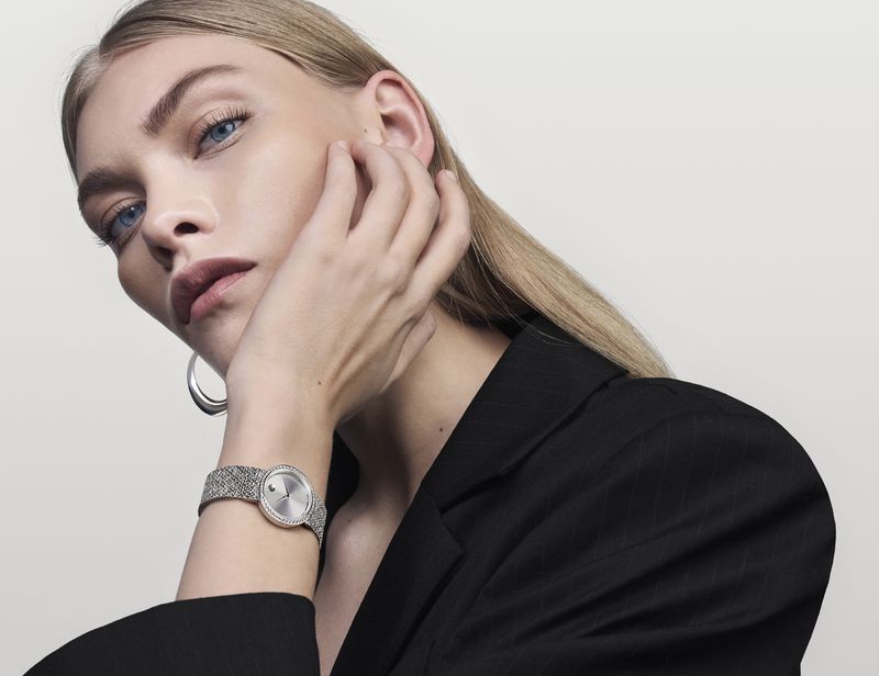Blonde woman in black blazer poses wearing silver wristwatch and hoop earring against a neutral background in a studio shot.