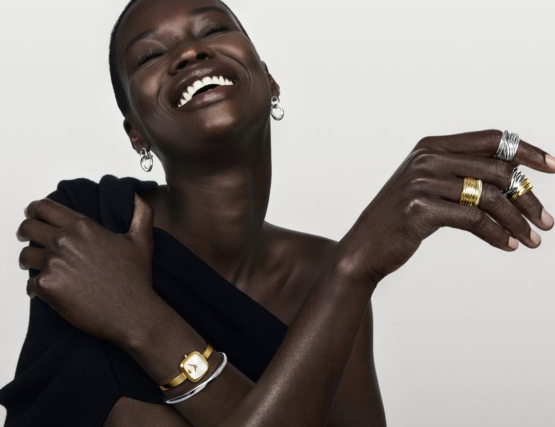 Smiling African woman with a bare shoulder poses in a close-up studio shot. The model wears various rings, earrings, and a gold watch.