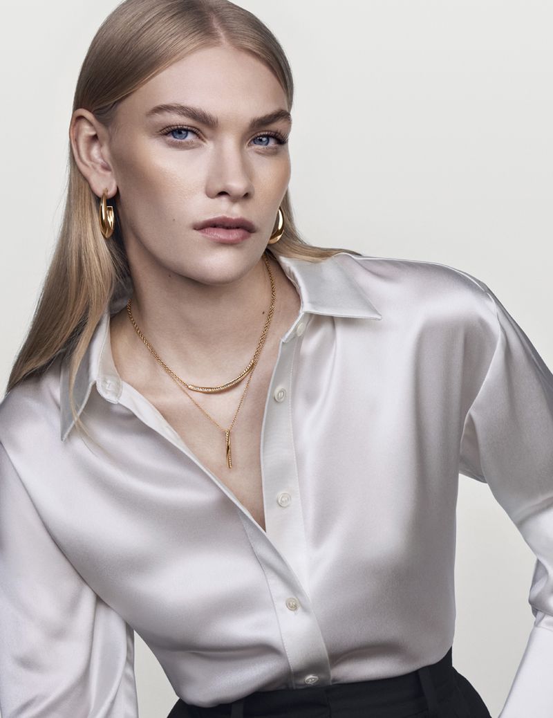 A fair-skinned adult woman with blond hair wears a white button-down shirt and gold jewelry against a neutral backdrop in a studio shot.