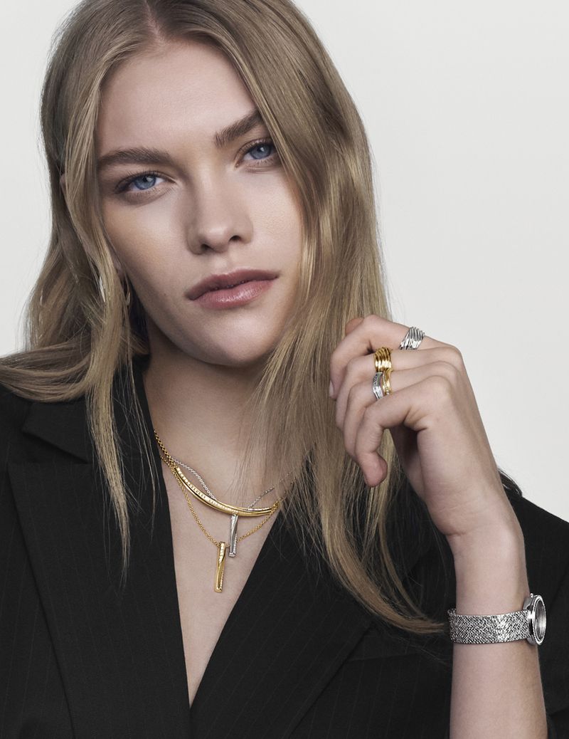 Close-up shot of a woman with blonde hair wearing rings, a necklace, and a watch, posing in a black blazer against a white background.