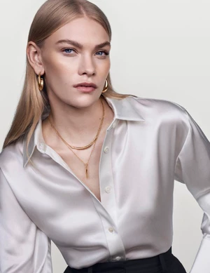 Model with blonde hair wearing a white silk shirt and gold necklaces against a neutral seamless studio background.