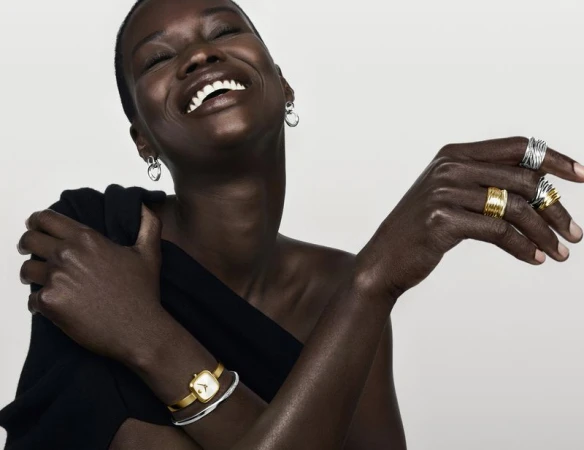 Smiling Black model wearing gold watch, silver rings, and earrings against a white background for Movado campaign