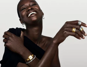 Smiling Black model wearing gold watch, silver rings, and earrings against a white background for Movado campaign