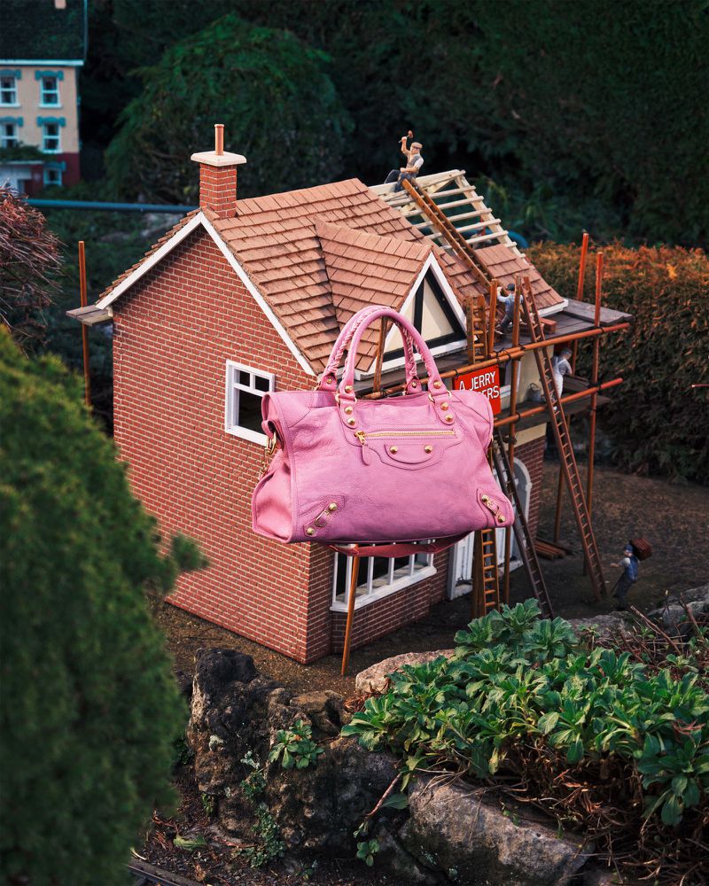 Pink leather tote bag floating near miniature brick house with construction workers on roof in detailed, outdoor diorama setting.