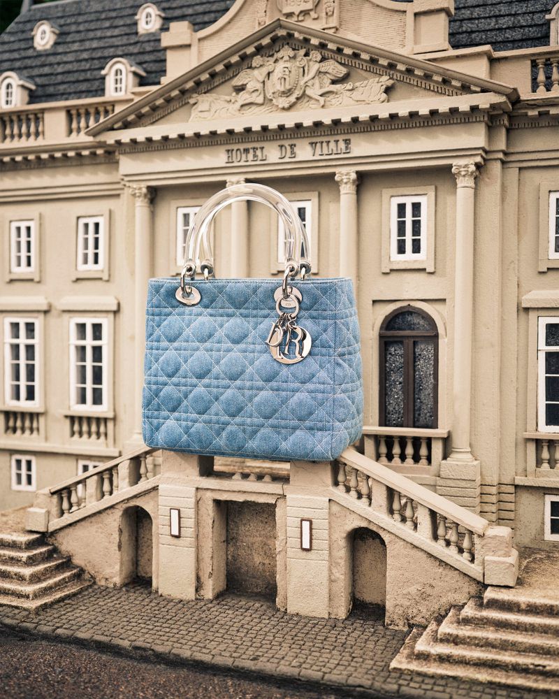 Elegant Lady Dior handbag in light blue quilted fabric with clear handle, displayed against a miniature building facade, fashion forward.