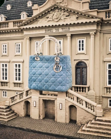 Elegant Lady Dior handbag in light blue quilted fabric with clear handle, displayed against a miniature building facade, fashion forward.