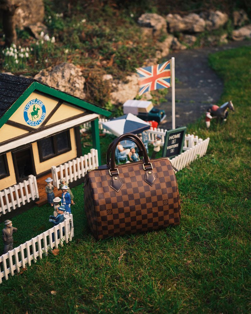 Brown checkerboard handbag on grass beside a miniature polo club with tiny figures, picket fence, British flag and stone path.