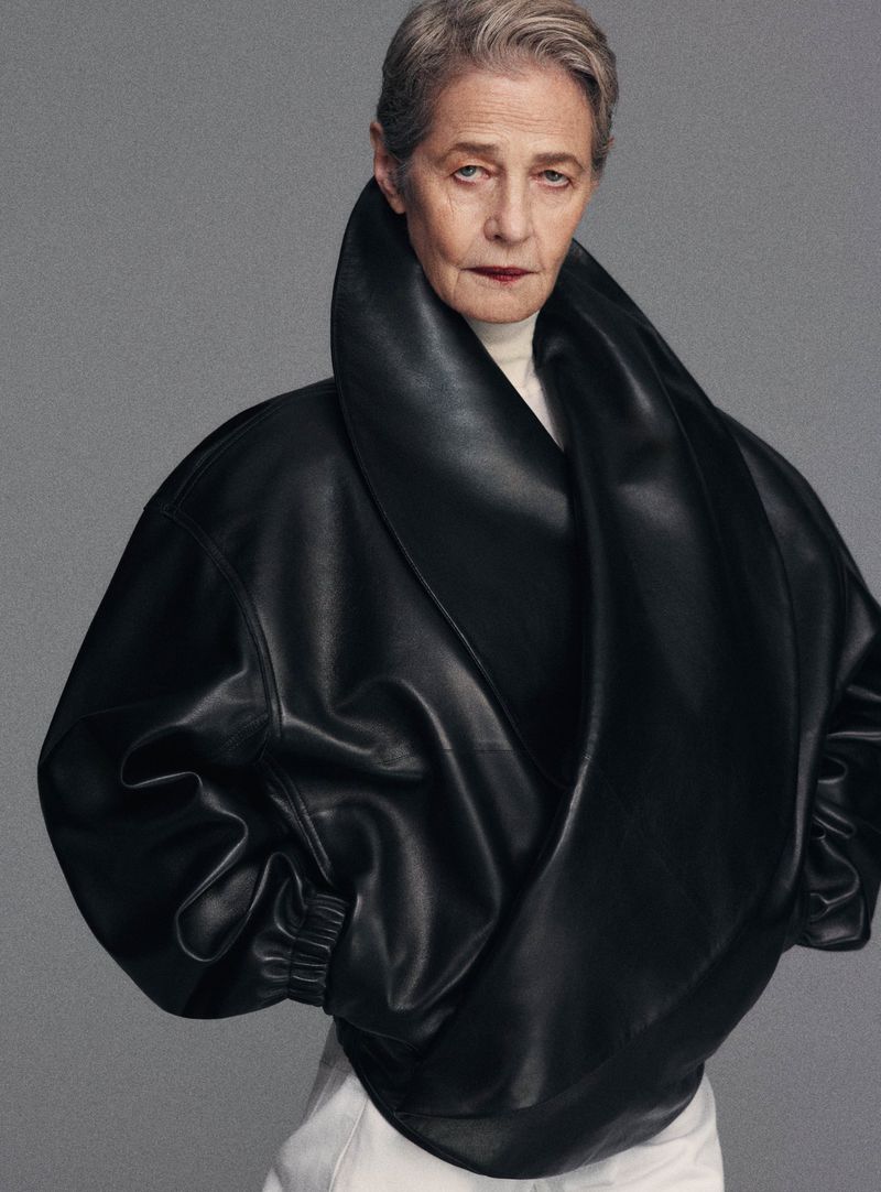 An older woman with short gray hair poses in a studio wearing a large, draped black leather jacket and white pants against a gray backdrop.