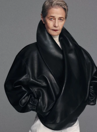 An older woman with short gray hair poses in a studio wearing a large, draped black leather jacket and white pants against a gray backdrop.
