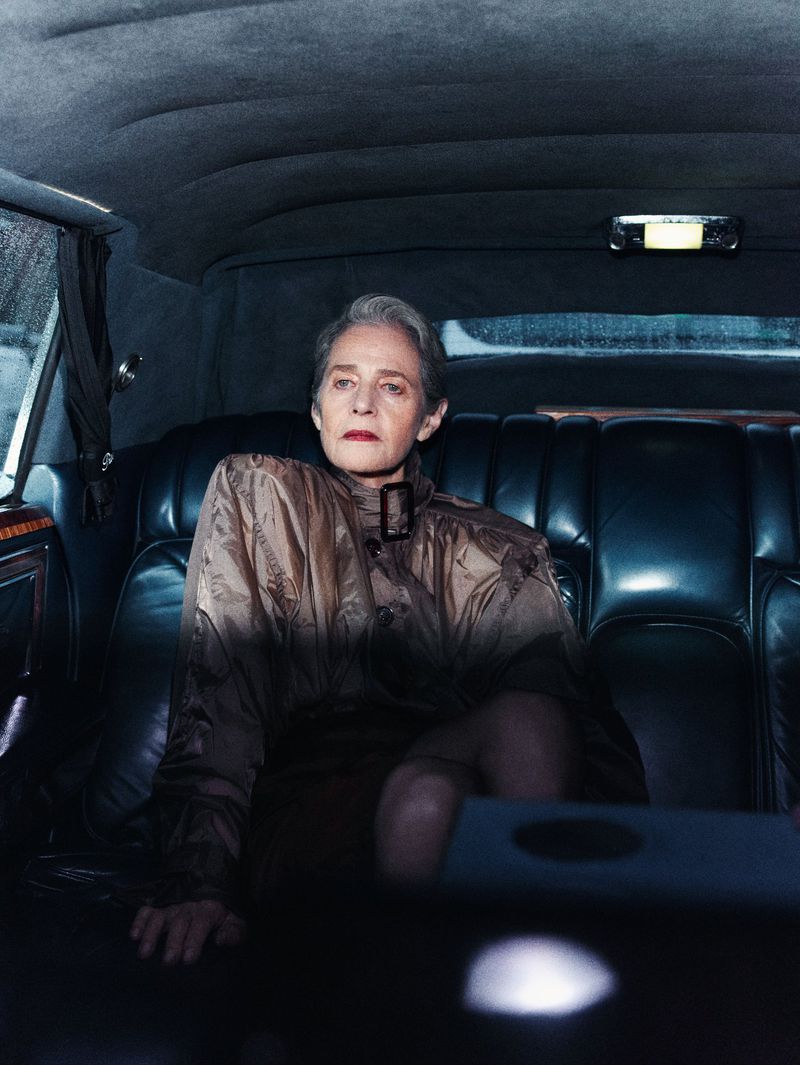 Older woman with short grey hair sits in the backseat of a limousine, wearing a patterned collared jacket, with dark interior details.