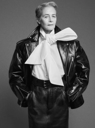 Elderly woman in black leather jacket and skirt poses in a studio with a neutral gray background, showcasing fashion and style.