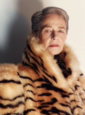 Portrait of an older woman with short gray hair wearing a tiger-striped fur coat, shot in a photo studio with neutral background.