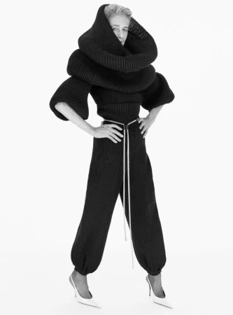 Woman in sculptural black knitwear stands in a studio, hands on hips, white stiletto heels. Monochrome fashion shot on white background.