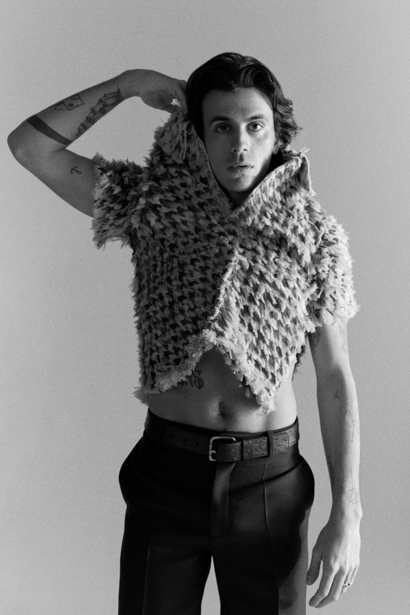 Black and white portrait of a young adult male posing indoors wearing a textured vest, dark pants, and a patterned belt against a grey background.