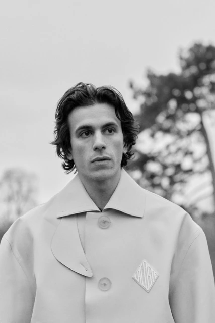 Black and white portrait of a young adult male in an overcoat against a blurred outdoor background of trees and cloudy sky.