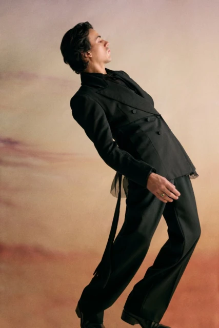 Young adult man poses in a black double-breasted suit, angled against a diffused sky background, displaying modern formal fashion.