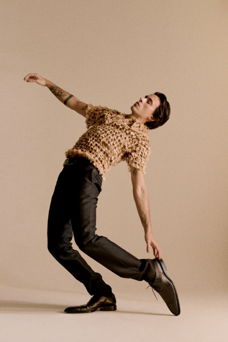 A male dancer poses in a knitted shirt and dark pants. He is leaning backwards with arms extended in an indoor studio setting.