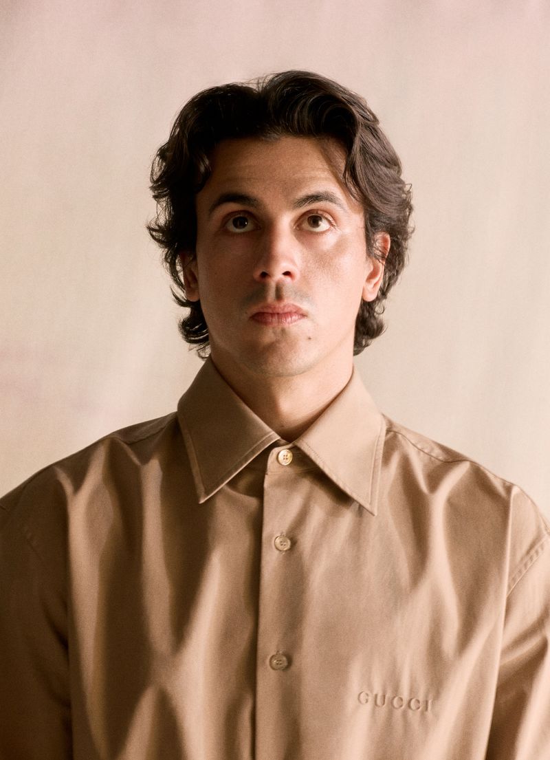 Portrait of a young adult male with dark hair wearing a tan button-down shirt with 'Gucci' logo, looking upward in a softly lit studio.