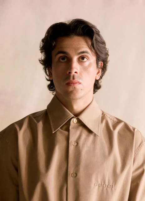 Portrait of a young adult male with dark hair wearing a tan button-down shirt with 'Gucci' logo, looking upward in a softly lit studio.