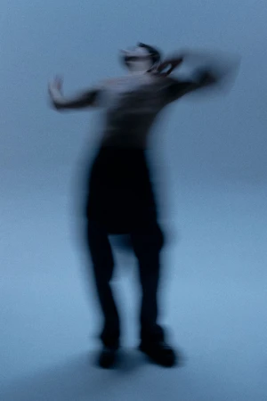 Blurred motion image of a person wearing dark pants and no shirt, dancing or moving expressively against a soft, light blue background.