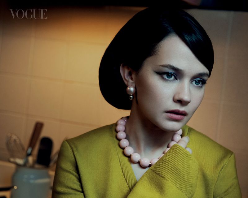 Portrait of a stylish woman with dark hair and pearl earrings wearing a chartreuse sweater and pink beaded necklace looking into the distance.