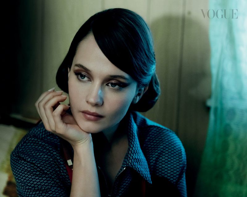 Woman with dark hair and flawless makeup poses elegantly indoors wearing a patterned shirt with a concerned facial expression and the Vogue logo.