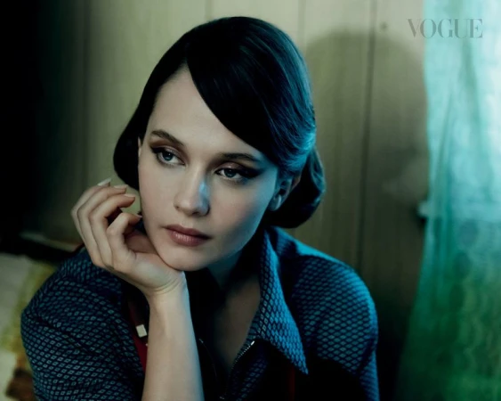 Woman with dark hair and flawless makeup poses elegantly indoors wearing a patterned shirt with a concerned facial expression and the Vogue logo.