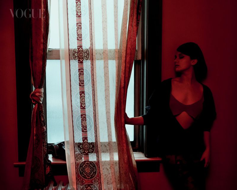 Woman in bra and cardigan stands by window with patterned curtains in a dimly lit room with red walls; Vogue logo visible in upper left.