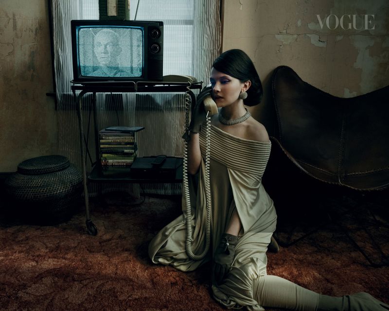 A woman in an off-shoulder dress and gloves kneels and speaks into a vintage telephone next to an old television set in a dark room.